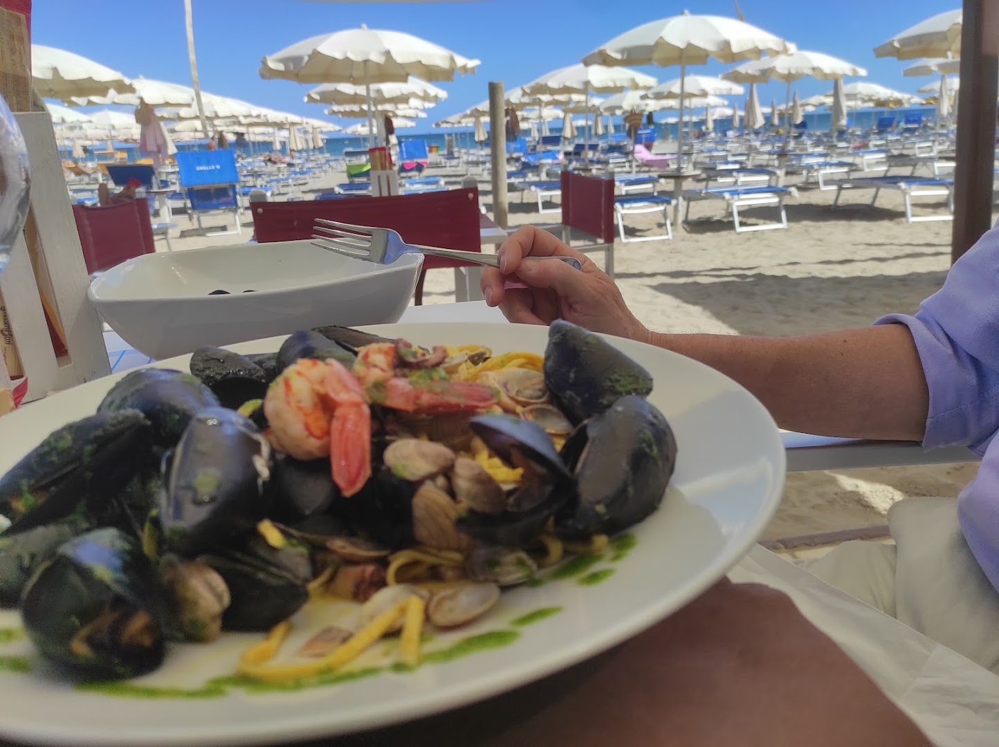 Piatto di tagliolini allo scoglio tenuto sopra un tavolo del Bagno Ettore, con la spiaggia e gli ombrelloni bianchi sullo sfondo