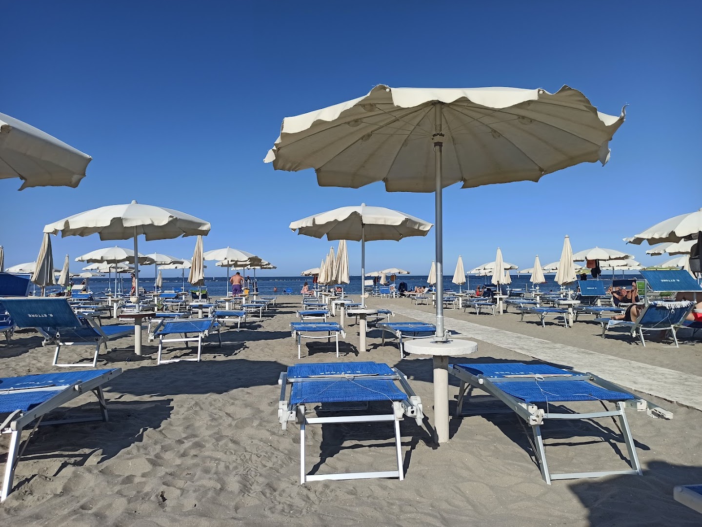 Ombrelloni bianchi aperti e lettini blu allineati sulla spiaggia del Bagno Ettore, mare in fondo